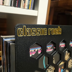 May include: Black vinyl record holder with the words "Classic Rock" in gold lettering. The holder has a honeycomb design with visible album covers. A shelf of CDs is visible in the background.