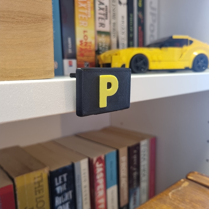 May include: A black plastic shelf label with a yellow letter "P" on a white bookshelf. A yellow toy car is on the shelf above the label.