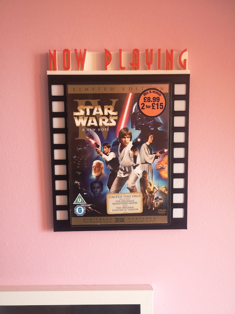 May include: A black framed movie poster with a film strip border. The poster features the Star Wars IV: A New Hope movie cover art. The poster is displayed on a pink wall with a sign that reads "NOW PLAYING".