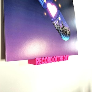 May include: A pink 3D printed record holder with the text "RECORD OF THE DAY" on it. The holder is attached to a wall and a record is displayed on top of it. The record features a colorful image of a heart and a city skyline inside a glass tube.