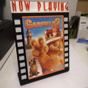 K&ouml;nnte beinhalten: Ein schwarz-wei&szlig;er Filmrollenst&auml;nder mit einem DVD-Geh&auml;use des Films "Garfield 2" im Inneren. Das DVD-Geh&auml;use zeigt ein Bild von Garfield der Katze, der eine Krone tr&auml;gt, und einem Hund, der ihn ansieht. Der St&auml;nder tr&auml;gt oben die Aufschrift "NOW PLAYING".