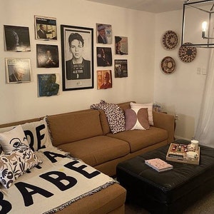 May include: A brown sectional sofa with a throw blanket that says "Made Babe" in black letters. The sofa is in a living room with a black coffee table and a wall decorated with framed record covers and woven wall hangings.