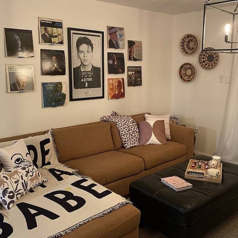 May include: A brown sectional sofa with a throw blanket that says "Made Babe" in black letters. The sofa is in a living room with a black coffee table and a wall decorated with framed record covers and woven wall hangings.