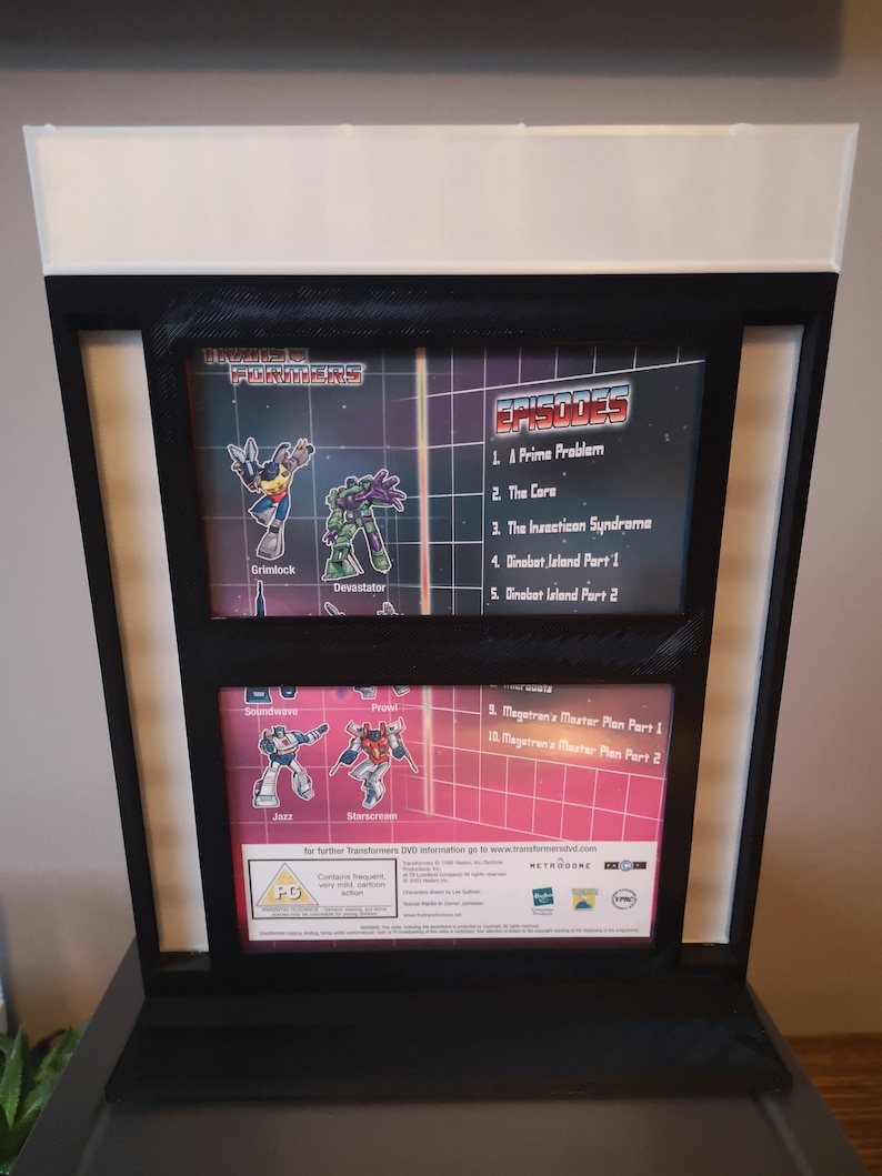 K&ouml;nnte beinhalten: Ein Transformers DVD-Geh&auml;use mit schwarzem und rotem Hintergrund. Die Vorderseite des Geh&auml;uses zeigt Bilder der Charaktere Grimlock, Devastator, Soundwave, Prowl, Jazz und Starscream. Das Geh&auml;use enth&auml;lt auch eine Liste der Episoden und eine Altersfreigabe von PG.