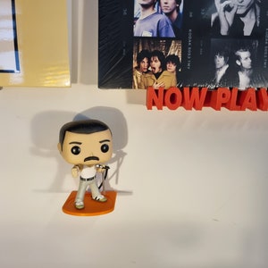 May include: A Funko Pop figure of Freddie Mercury, the lead singer of Queen, wearing a white tank top, black belt, and gray pants. He is holding a microphone and standing on an orange base. The figure is on a white wall with a red sign that says "NOW PLAY" and a framed photo of a band behind it.