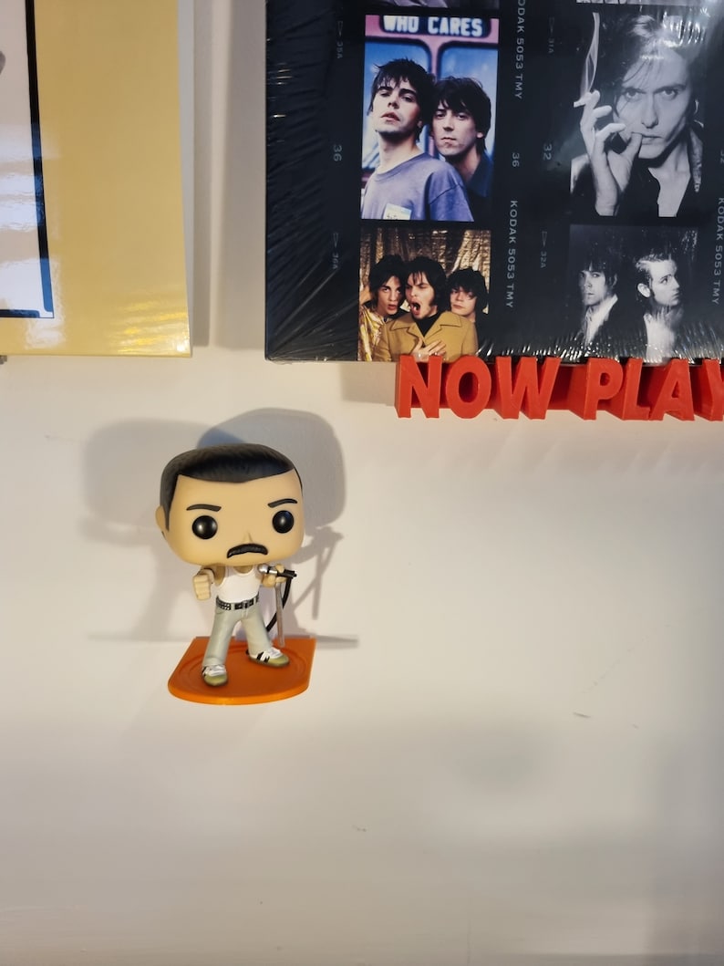 May include: A Funko Pop figure of Freddie Mercury, the lead singer of Queen, wearing a white tank top, black belt, and gray pants. He is holding a microphone and standing on an orange base. The figure is on a white wall with a red sign that says "NOW PLAY" and a framed photo of a band behind it.