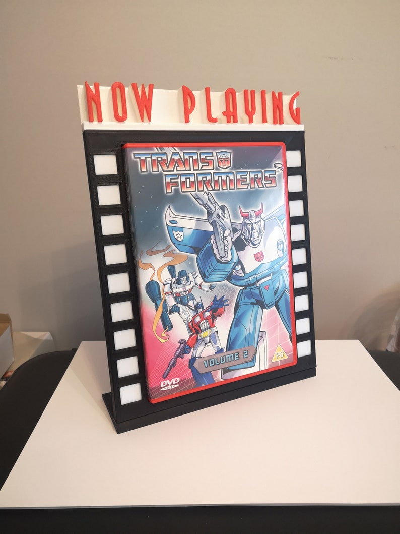 K&ouml;nnte beinhalten: Ein 3D-gedrucktes Kinoplakat mit schwarzem Rahmen und rotem "NOW PLAYING"-Schild. Das Plakat zeigt ein DVD-Geh&auml;use f&uuml;r "Transformers Volume 2".