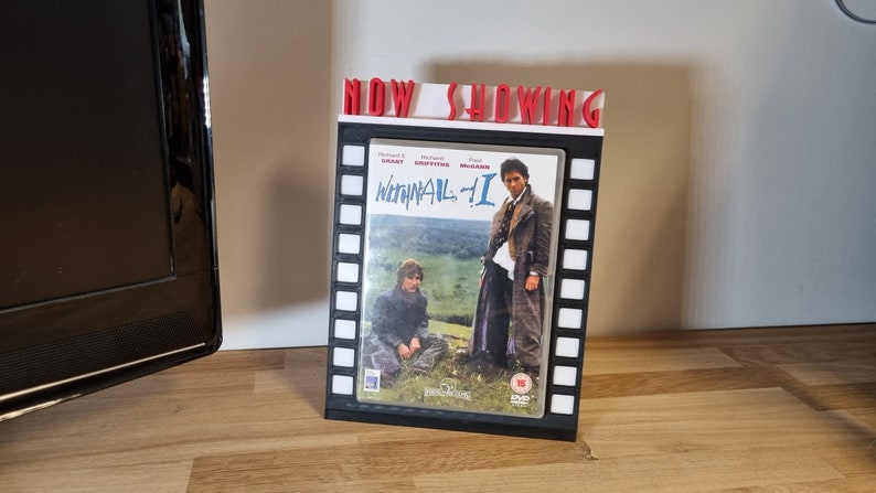 K&ouml;nnte beinhalten: Ein schwarz-wei&szlig;er Filmrollenst&auml;nder mit einem roten Schild "NOW SHOWING". Der St&auml;nder h&auml;lt ein DVD-Geh&auml;use mit dem Film "Withnail & I" darauf.