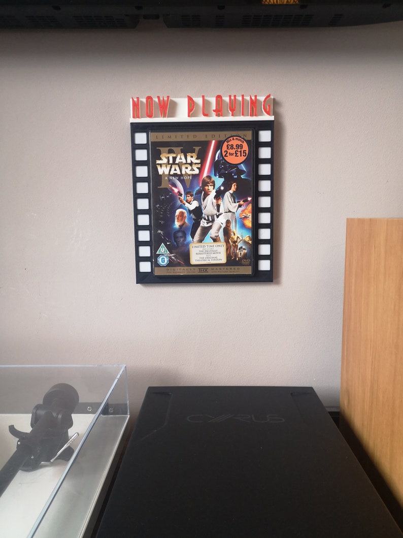 May include: A white and red "Now Playing" sign with a black and white movie poster of Star Wars: A New Hope. The poster is in a frame that looks like a film reel. The poster has a red price tag that says "&pound;8.99 2 for &pound;15".