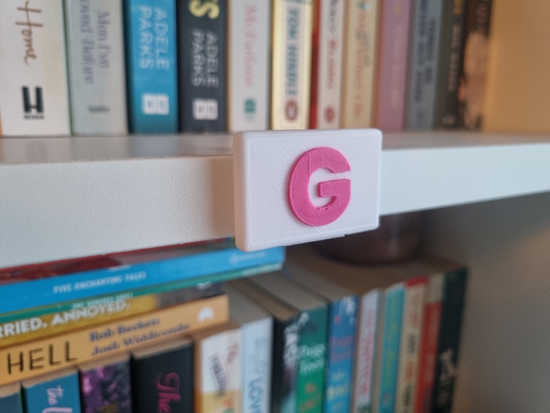 May include: A white shelf with a pink letter G on a white label attached to the shelf.