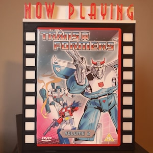 K&ouml;nnte beinhalten: Ein schwarz-wei&szlig;es Kinoplakat mit roter Aufschrift "NOW PLAYING" und einem DVD-Geh&auml;use mit einer rot-blau-wei&szlig;en Illustration von Transformers-Charakteren. Das DVD-Geh&auml;use tr&auml;gt den Titel "Transformers" und "Volume 2" aufgedruckt.
