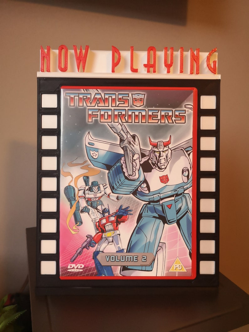 K&ouml;nnte beinhalten: Ein schwarz-wei&szlig;es Kinoplakat mit roter Aufschrift "NOW PLAYING" und einem DVD-Geh&auml;use mit einer rot-blau-wei&szlig;en Illustration von Transformers-Charakteren. Das DVD-Geh&auml;use tr&auml;gt den Titel "Transformers" und "Volume 2" aufgedruckt.