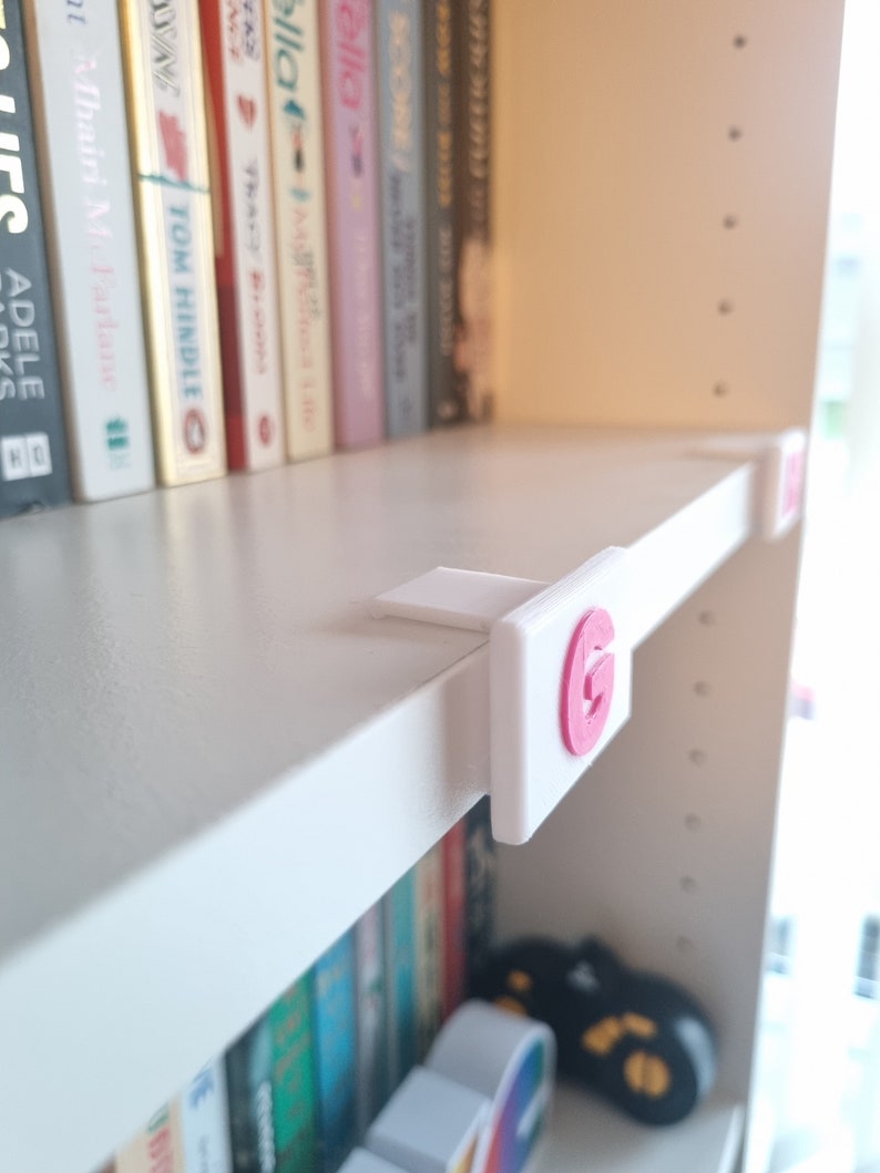 May include: White bookshelf with a pink letter G on a white label attached to the shelf. The letter G is on a white background.
