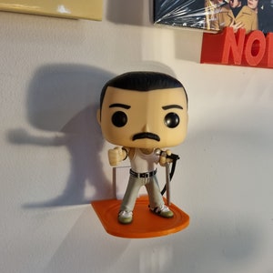 May include: A Freddie Mercury Funko Pop figure holding a microphone, standing on an orange plastic shelf. The figure is wearing a white tank top, black belt, and grey pants. The figure has black hair, a mustache, and a serious expression.