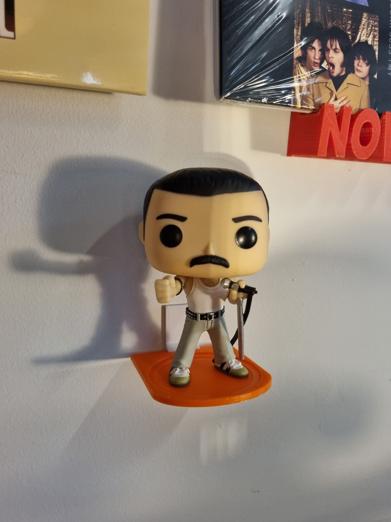 May include: A Freddie Mercury Funko Pop figure holding a microphone, standing on an orange plastic shelf. The figure is wearing a white tank top, black belt, and grey pants. The figure has black hair, a mustache, and a serious expression.
