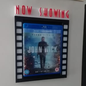 May include: A black frame with a white border and the words "NOW SHOWING" in red letters. The frame holds a Blu-ray disc case with a movie poster for "John Wick" featuring Keanu Reeves. The poster has a blue background with a black and white image of Keanu Reeves holding a gun. The poster also has the words "John Wick" in white letters and the words "Don't Get Him Off" in smaller white letters.