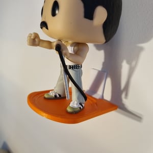 May include: A small orange plastic shelf with a white backing, designed to hold a figurine. A Freddie Mercury Pop figure is standing on the shelf, holding a microphone.