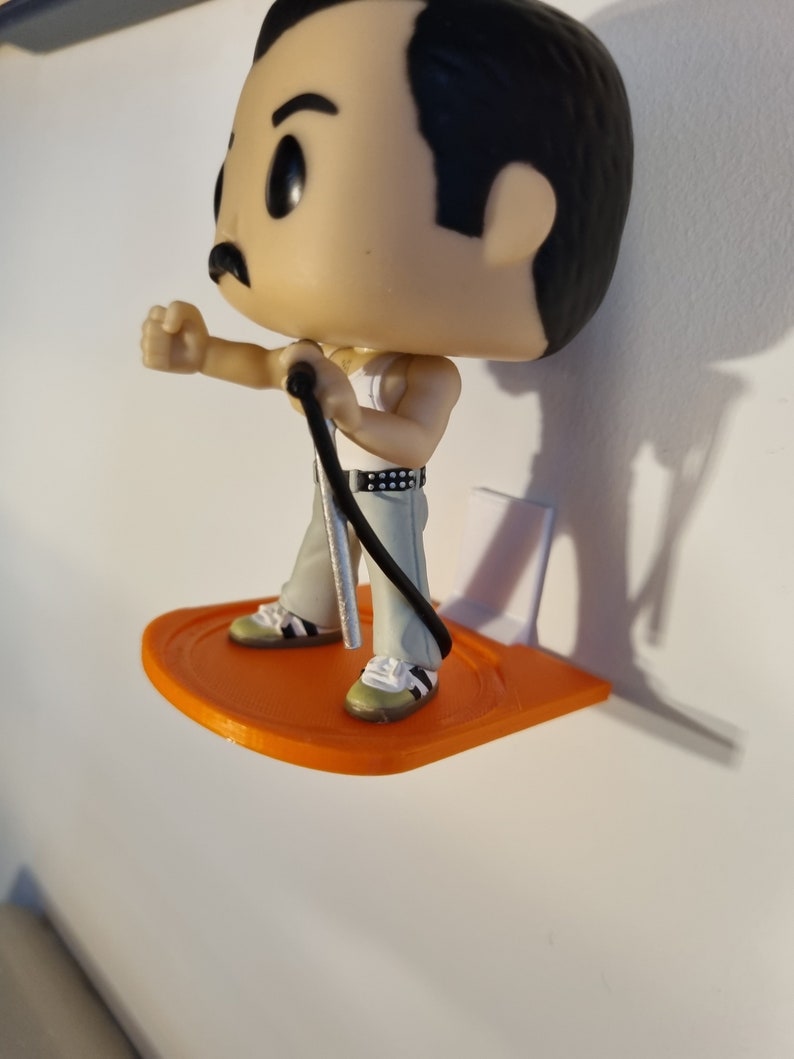 May include: A small orange plastic shelf with a white backing, designed to hold a figurine. A Freddie Mercury Pop figure is standing on the shelf, holding a microphone.
