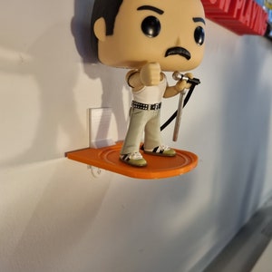 May include: A figurine of Freddie Mercury, the lead singer of Queen, holding a microphone. The figurine is standing on an orange shelf attached to a white wall. The shelf is labeled "NOW PLAYING" in red letters.