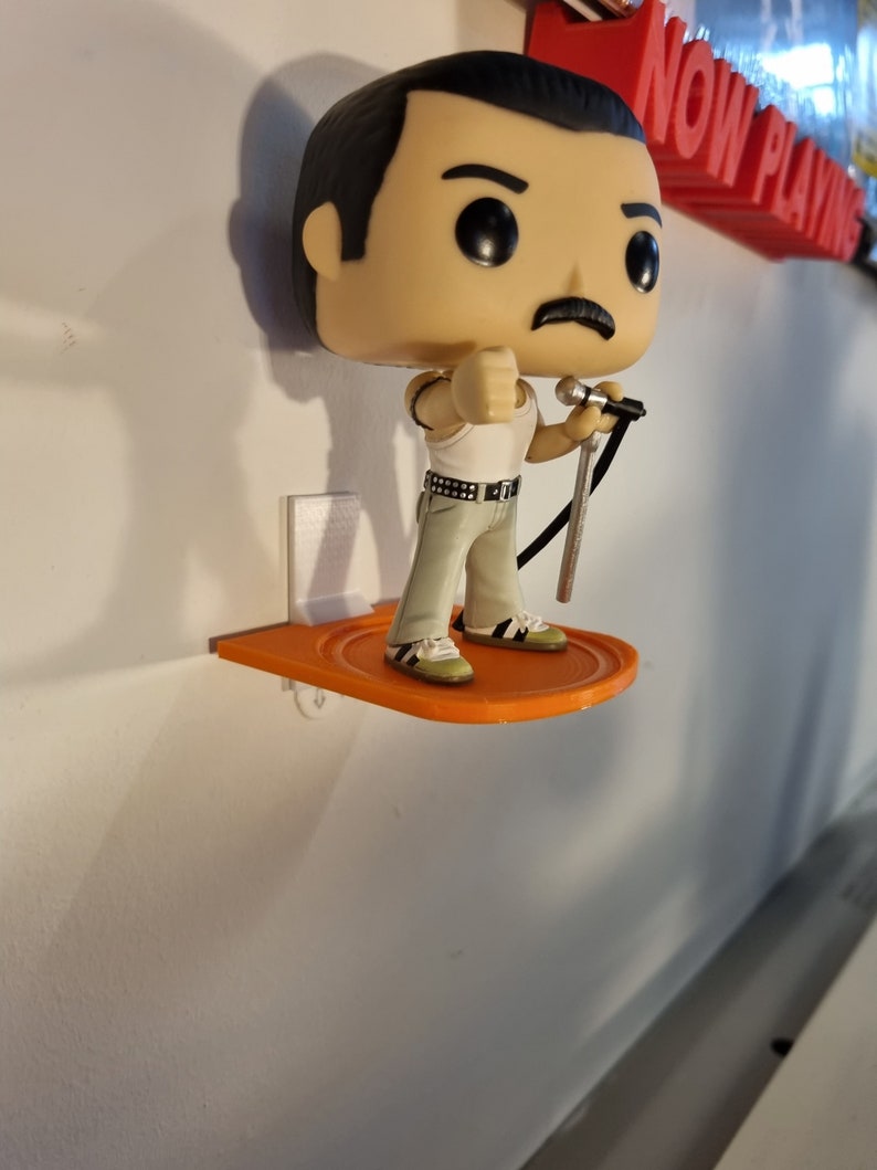 May include: A figurine of Freddie Mercury, the lead singer of Queen, holding a microphone. The figurine is standing on an orange shelf attached to a white wall. The shelf is labeled "NOW PLAYING" in red letters.