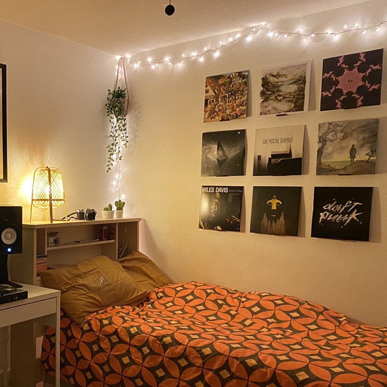A bedroom with a display made up of a square of nine vinyl records on display shelves above the bed. There is a row of fairy lights hung along the top near the ceiling and a plant hangs in the corner.