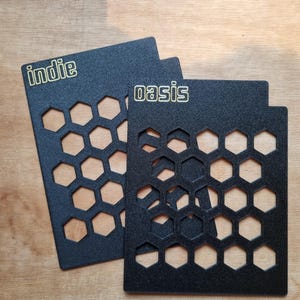 May include: Two rectangular black metal stencils featuring a repeating honeycomb pattern of hexagonal cutouts. The left stencil has the word indie printed in gold, and the right stencil has the word oasis printed in gold at the top.