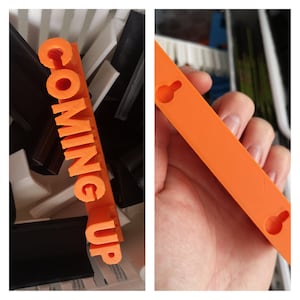 Może przedstawiać: Pomarańczowy znak wydrukowany w 3D z napisem „COMING UP” pogrubioną czcionką. Znak jest prostokątny i ma teksturowaną powierzchnię. Na zdjęciu widać również inne obiekty drukowane w 3D w kolorze czarnym, białym i szarym.