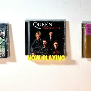 May include: A wall display of three music CDs. The middle CD is a Queen Greatest Hits album with a photo of the band members. The text "NOW PLAYING" is displayed below the CD. The CD on the left is a Restraining Bolt album. The CD on the right is a Cream Anthems 2001 album.