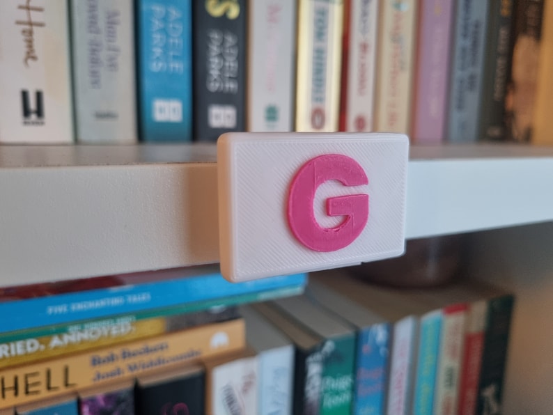May include: A white shelf with a pink letter G on a white rectangular label. The shelf is filled with books.
