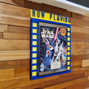 May include: A blue and yellow framed movie display with the words "NOW PLAYING" at the top. The display features a Star Wars movie case. The case has a blue background with images of characters and the title "Star Wars: A New Hope".