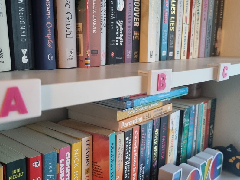 May include: A white bookshelf with books organized by letter. The top shelf has books with titles like "The Love Hypothesis" and "Malibu Rising". The bottom shelf has books with titles like "Lessons in Chemistry" and "Nick Hornby".