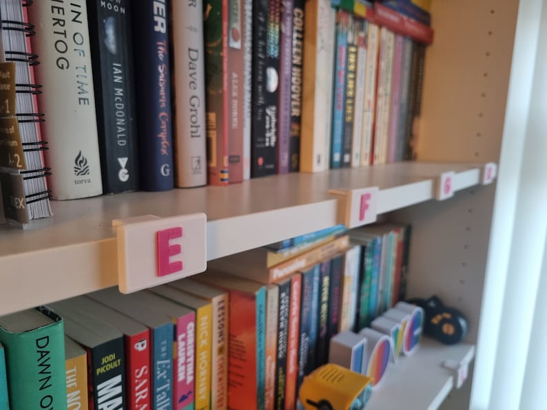 May include: A bookshelf with books organized by letter. White plastic dividers with pink letters E, F, G, and D are attached to the shelves.