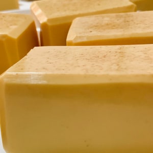 May include: Close-up of several rectangular bars of soap. The soap is a light yellow colour with a slightly textured surface and a dusting of brown particles. The bars have a smooth, glossy finish and are arranged closely together.