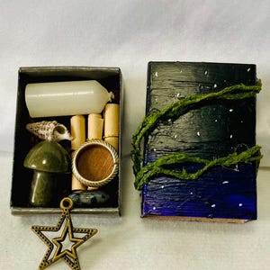 Könnte beinhalten: Eine kleine, offene Metallbox mit einer weißen Kerze, einer Muschel, einem grünen Pilz, einem Ring und kleinen Schriftrollen. Daneben eine kleine, geschlossene Box, bemalt mit einem Nachthimmel-Design und mit grünen Ranken verziert. Ein bronzener Sternanhänger ist davor.