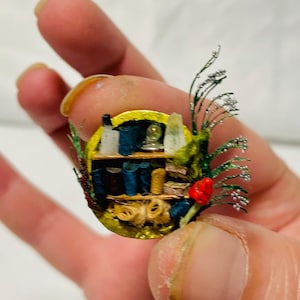 May include: A miniature diorama of a bookshelf with tiny books, bottles, and decorative items. The circular frame has a yellow border, green and silver accents, and a red flower. The diorama is held in a hand.