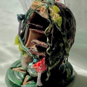 May include: A handcrafted miniature fairy house sculpture. The dark brown structure is decorated with green vines, red berries, and a red and white mushroom. It rests on a green leaf-shaped base, creating a fantasy theme.