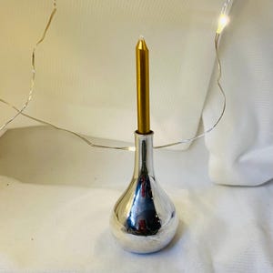 Peut inclure: Un bougeoir argenté avec une bougie dorée. Le support a une base arrondie et un col étroit. Une bougie dorée est insérée en haut. Le fond est blanc avec une guirlande lumineuse.