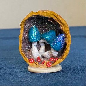 May include: A miniature scene of blue mushrooms with white stems growing inside a halved walnut shell. The inside of the shell is decorated with glitter and red dots.