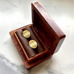 Puede incluir: Un par de gemelos dorados con líneas horizontales, exhibidos en una caja de madera oscura abierta con interior marrón. La caja tiene bisagras y un acabado pulido, lo que sugiere una joya o un accesorio.