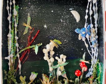 May include: A small diorama depicting a night scene with a crescent moon and stars. The artwork includes handmade plants, mushrooms, and abstract shapes in green, red, white, and blue, all contained within a white-framed box.