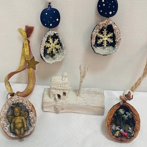 Könnte beinhalten: Eine Sammlung handgefertigter Weihnachtsschmuck. Die Ornamente umfassen Walnussschalen-Designs mit Engel-, Schneeflocken- und Miniatur-Winterszenen. Die Ornamente sind mit goldenen, weißen und blauen Akzenten verziert und werden mit Bändern und Schnüren aufgehängt.
