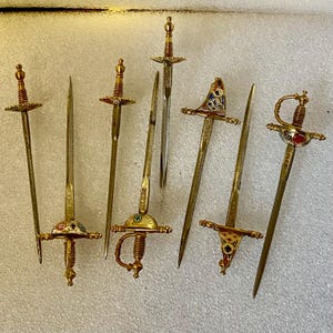 Könnte beinhalten: Eine Sammlung von dekorativen Miniaturschwertern mit verzierten goldfarbenen Griffen und Klingen. Die Schwerter weisen verschiedene Designs auf, darunter detaillierte Parierstangen und Schmuckakzente. Die Klingen sind silber- und goldfarben, und die Schwerter variieren in Größe und Stil.