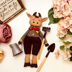 May include: A brown and white stuffed bull with a red and white checkered shirt, a red bow tie, and a bell. The bull is holding a small black and brown gardening tool. The bull is sitting on a white surface with a green plant and a sign that says "Remember you decide to be yourself".