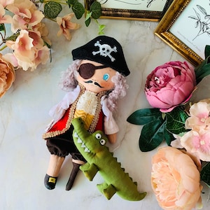 Pode incluir: Uma boneca pirata feita à mão com um tapa-olho preto, uma jaqueta vermelha e dourada e um chapéu de pirata preto com um design de caveira e ossos cruzados. A boneca está segurando um brinquedo de jacaré verde.