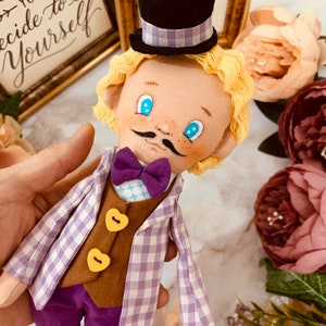 May include: A handmade doll with a whimsical design. The doll has a black top hat with a purple checkered band, a purple bow tie, and a checkered purple and white jacket. The doll's vest has two yellow heart-shaped buttons. The doll has blonde hair, blue eyes, and a black mustache.