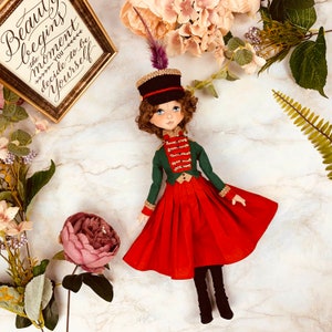 May include: A handmade doll wearing a green and gold military-style jacket with a red skirt and black boots. The doll has brown hair and blue eyes and is wearing a black hat with a feather.
