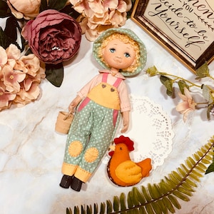 May include: A handmade doll with blonde hair wearing a green and yellow polka dot jumpsuit and a yellow hat. The doll is holding a small bucket and is standing next to a yellow felt chicken.