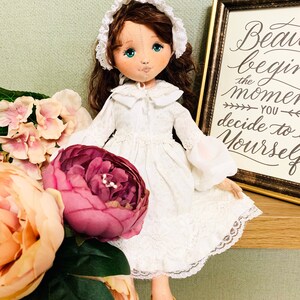 May include: A white fabric doll with brown hair wearing a white dress and a white bonnet. The doll is sitting on a shelf with a sign that reads "Beauty begins the moment you decide to be yourself."