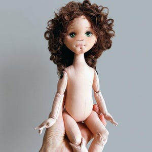 May include: A handcrafted fabric doll with a pale complexion, freckles, and vibrant green eyes. The doll features voluminous, curly dark brown hair and articulated limbs connected by small black buttons at the shoulders and hips.