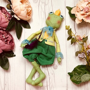 Peut inclure: Une poupée de grenouille verte portant une veste jaune avec un col en dentelle bleu et blanc et un pantalon vert. La poupée porte une couronne et a une plume attachée à sa veste.